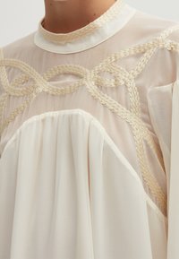 Light beige blouse with a sheer fabric, featuring a high collar and intricate braided detailing across the chest. Soft texture, flowing design.