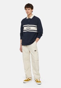 Marineblauwe polo met lange mouwen, voorzien van een witte streep en logo, gecombineerd met crèmekleurige cargobroeken en mosterdkleurige sneakers, met een relaxte pasvorm en knoopkraag.
