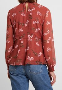 Kvinde iført en rustfarvet bluse med pink blomsterprint og lange ærmer, parret med blå jeans, vist fra ryggen.