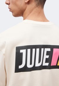 Άνδρας με μπεζ πουκάμισο με τη λέξη «JUVE» σε μεγάλα λευκά γράμματα σε μαύρο ορθογώνιο, με ροζ και κίτρινα μπλοκ δεξιά.