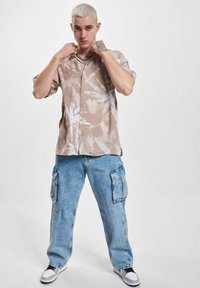 Kurzärmeliges Hemd in Beige mit weißen abstrakten Blättern, kombiniert mit hellblauen Cargo-Jeans mit seitlichen Taschen und entspannter Passform.