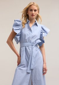 Rochie bleu rayată cu mâneci scurte și volănașe, cu nasturi pe față și un cordon la talie. Țesătură fină cu croială lejeră și buzunare laterale.