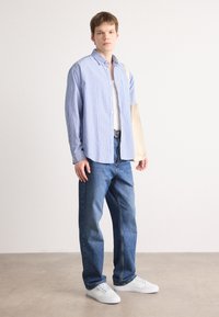 Un jeune homme se tient debout, portant une chemise à rayures bleues, un t-shirt blanc, un jean bleu, des baskets blanches et portant un sac fourre-tout beige sur l'épaule.