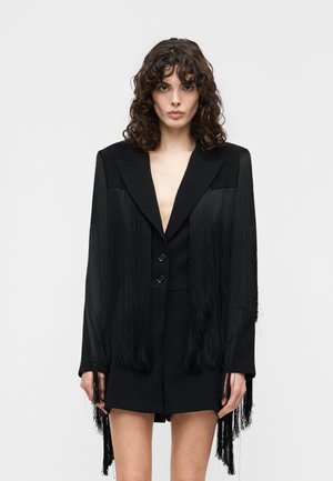 Kobieta o kręconych włosach ubrana w czarną sukienkę-żakiet z długimi frędzlami na rękawach na jasnym, jednolitym tle.