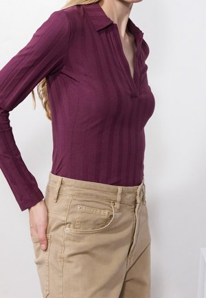 Vrouw draagt een aansluitend paars geribd shirt met lange mouwen en beige hoog getailleerde broek, hand in de zak tegen een effen achtergrond.