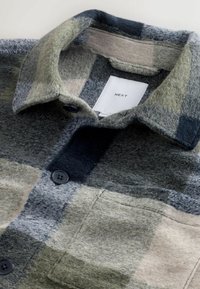 Wool shirt in shades of green, gray, and black with a plaid pattern. Features a collar, black buttons, and a tag labeled "NEXT."