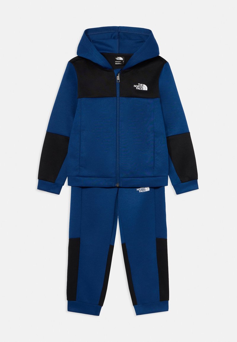 Modri dvodelni trenirka, ki vključuje jopico s kapuco na zadrgo z črnimi poudarki in ujemajoče se hlače, izdelana iz mehke tkanine z gladko teksturo.