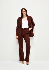 Bruine op maat gemaakte blazer met bijpassende broek, voorzien van een enkele knoop, zijzakken en een gladde textuur. Witte V-hals top eronder.
