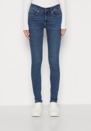 Slim-fit blauwe spijkerbroek met voorzakken en knoopsluiting, gedragen met witte sneakers en een wit langarmshirt tegen een effen achtergrond.
