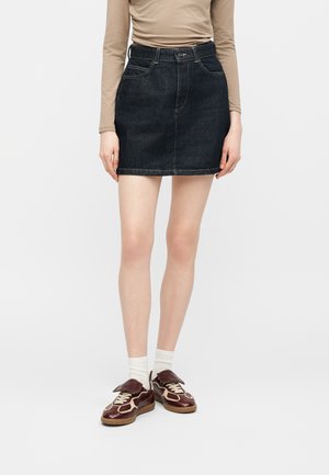 Femme portant un haut à manches longues beige, une mini-jupe en jean noire, des chaussettes blanches et des baskets marron et crème, debout devant un fond uni.