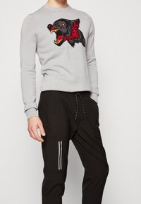 Person som bär en ljusgrå tröja med design av ett röd- och svartfärgat vrålande varghuvud, tillsammans med svarta byxor med dragsko och dragkedjeficka.