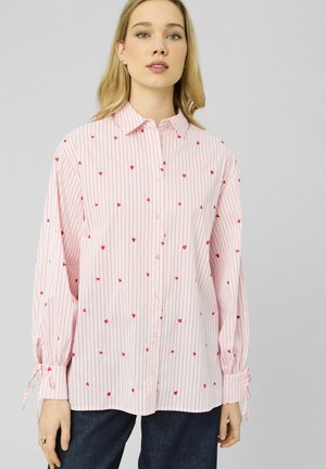 Femme portant une chemise légère rose clair à boutons, ornée de fines rayures rouges et de petits imprimés floraux rouges éparpillés, associée à un jean foncé.