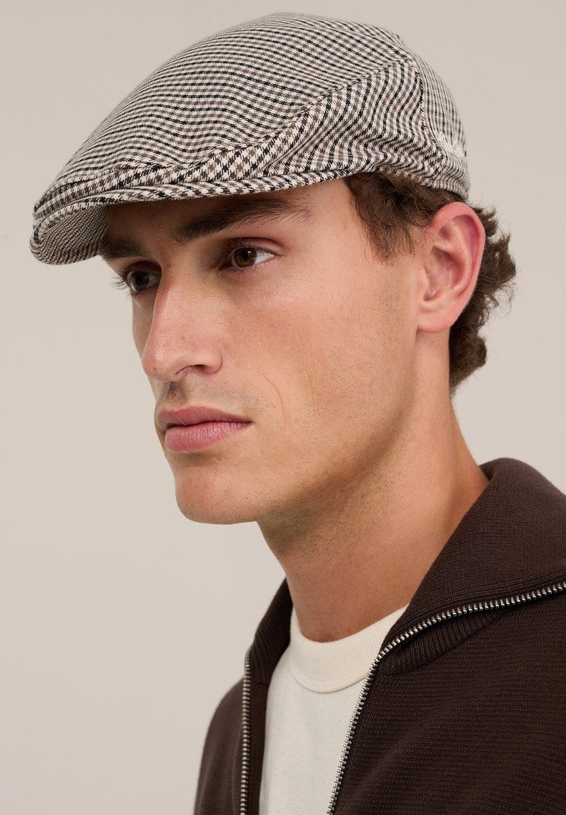 Jeune homme portant une veste marron zippée et une chemise blanche, regardant vers la gauche, avec une casquette plate à carreaux noir et blanc sur la tête.
