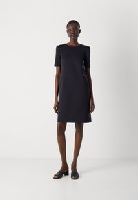 Robe navy à manches courtes avec un col arrondi, dotée de deux poches latérales et d'un léger éclat. Associée à des sandales noires à talons carrés ouvertes.