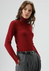 Rode coltrui van zachte stof met lange mouwen. Heeft zichtbare naden en knoopaccenten op de manchetten. Gecombineerd met grijze, hoogmidjse broek.