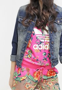 Kvinna som bär en blå denimjacka över en rosa blommig Adidas-tröja och matchande blommiga byxor med en rosa dragsko.
