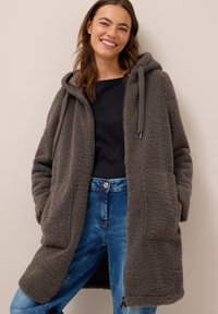 Manteau en polaire gris avec une capuche, présentant une fermeture éclair, deux poches avant et des cordons de serrage. Porté par-dessus un haut noir et un jean bleu.