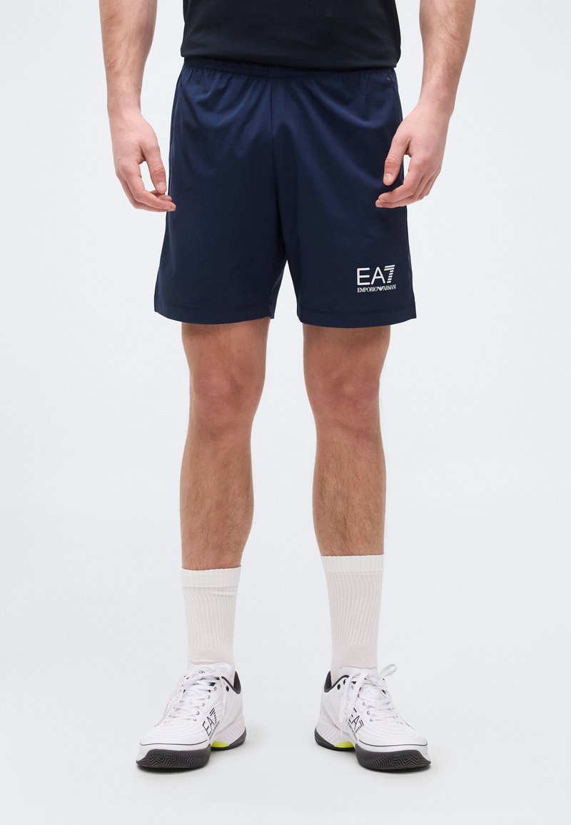 Άνδρας που φοράει σορτς EA7 Emporio Armani σε ναυτικό μπλε, λευκές κάλτσες και λευκά αθλητικά παπούτσια με το λογότυπο EA7, στέκεται μπροστά σε απλό φόντο.