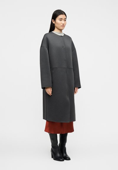 Γυναίκα που φοράει ένα μακρύ γκρι ανθρακοκάλυβα παλτό με κουμπιά πάνω από μια σκουριασμένη φούστα και μαύρες μπότες με τακούνια μέχρι το γόνατο, στέκεται μπροστά σε ένα απλό φόντο.