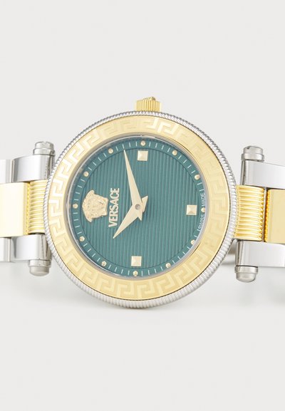 Gold and silver-toned watch with a teal textured dial, Greek key pattern on the bezel, and a lion head logo at 12 o'clock. Gold accents and square hour markers.