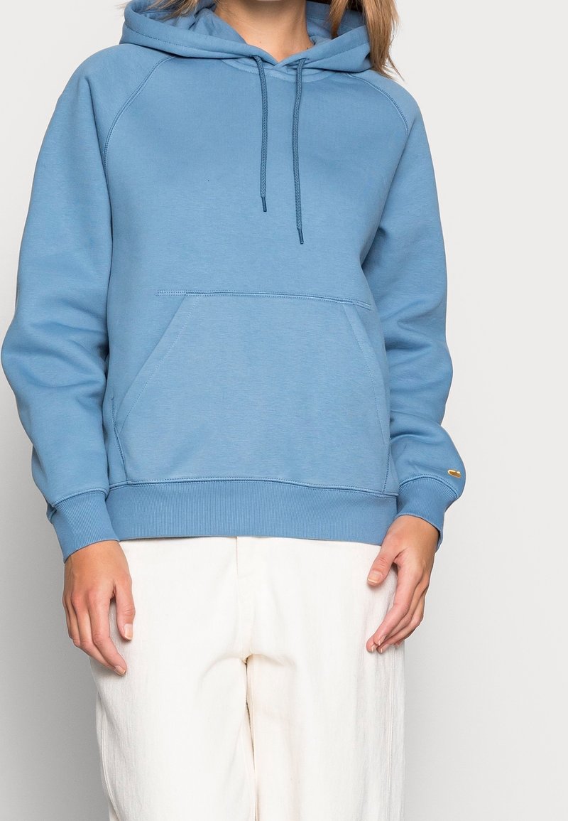 Donna che indossa un hoodie azzurra chiara con tasca frontale e pantaloni avorio, in piedi contro uno sfondo chiaro e uniforme.