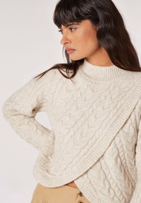 Beige kabelstrik sweater med høj ribbede krave og diagonalt design. Blød tekstur, lange ærmer og fremtrædende kabelmønster detaljer.