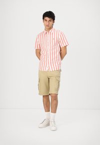 Maglietta a righe rosse e bianche a maniche corte, abbinata a pantaloni cargo beige e scarpe da ginnastica bianche. La maglietta presenta un design con bottoni.