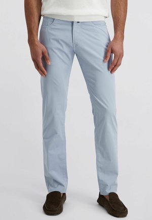 Mann mit hellblauen Hosen und dunkelbraunen Loafers, der vor einem einfarbigen weißen Hintergrund steht.