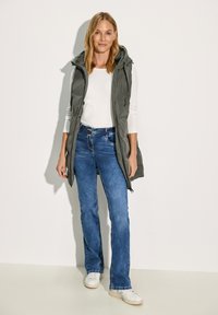 Puffige graue Weste mit Kapuze, weißes Langarmshirt darunter, blaue Schlagjeans und weiße Sneakers. Sanfte Texturen, funktionales Design.