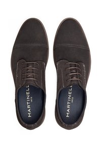 Zapatos de ante marrón con cordones y una textura suave, con acentos de costura blanca y una plantilla azul oscuro que muestra el nombre de la marca.