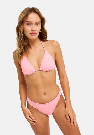 Kobieta z falującymi jasnobrązowymi włosami, ubrana w jasnoróżowy halter bikini top i pasujące do tego doły, stojąca na jasnym, jednolitym tle.