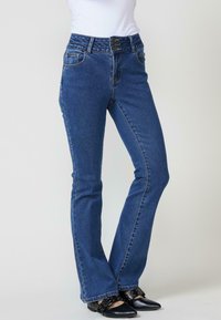 Jeans acampanados de mezclilla azul con cintura alta, cierre de botón único, cinco bolsillos y costuras en contraste. Combinados con zapatos planos negros.