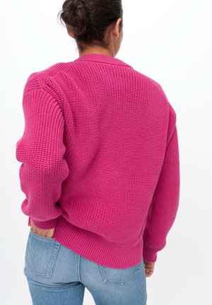 Pull en tricot rose avec un motif texturé, coupe oversize, poignets et ourlet côtelés, associé à un jean en denim bleu, vu de dos.