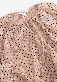 Blouse en chiffon rose avec un motif en dentelle, des pois noirs, des accents froncés et un décolleté en clé. Étiquette visible au col.