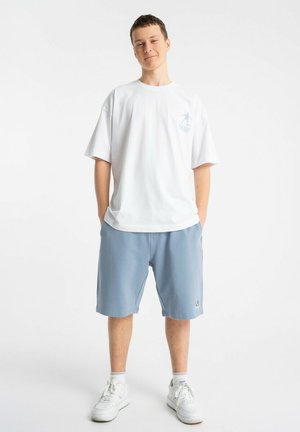 Młody mężczyzna stoi z rękami w kieszeniach, ubrany w białą, luźną koszulkę, jasnoniebieskie szorty, skarpetki i białe trampki na białym tle.