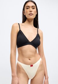 Bralette triangular negro con detalle de malla transparente y tirantes ajustables, combinado con tanga color crema con una etiqueta de logo roja.