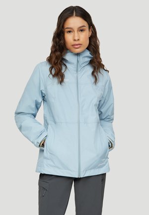 Femme portant une veste zippée bleu clair, les mains dans les poches, et un pantalon gris foncé, debout devant un fond uni.