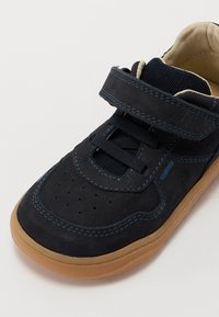 Marineblå fløyelslær-sneaker med borrelås, kontrasterende blå sømmer og såle i gummi. Har perforeringer for pusteevne.