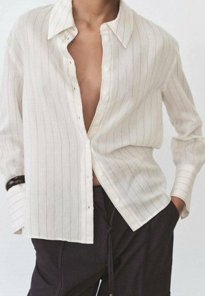 Osoba ubrana w białą, częściowo rozpiętą, koszulę z długim rękawem z cienkimi pionowymi paskami oraz czarne spodnie, z rękami w kieszeniach.