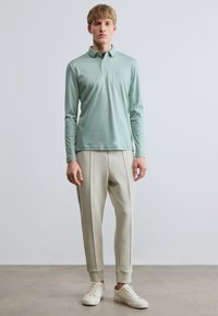 Világoszöld hosszú ujjú galléros póló, világos bézs színű, elasztikus mandzsettás és oldalsó varrással díszített jogger nadrággal párosítva. Fehér tornacipő.