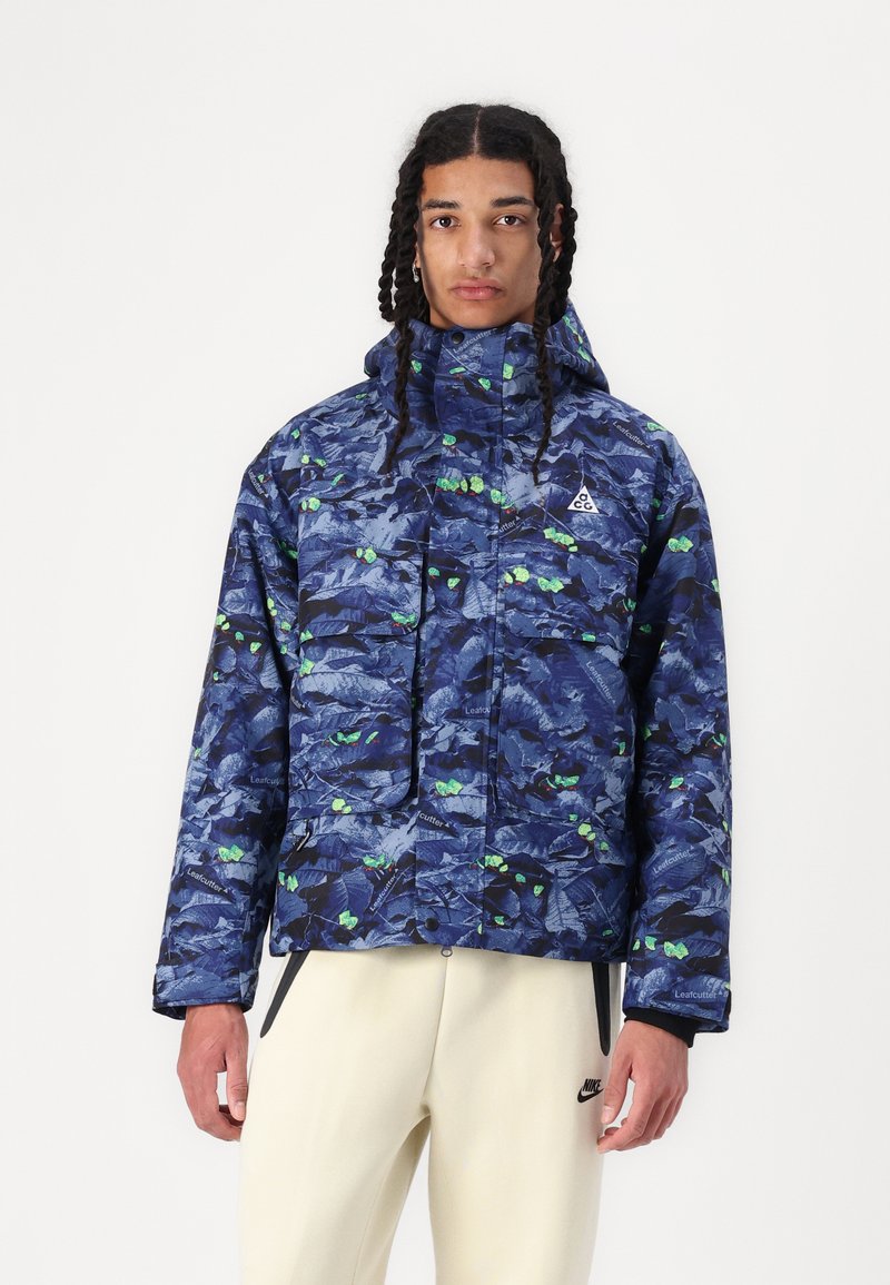 Blue camo-patterned puffer jacket with green accents, hood, and front pockets. Made of a smooth, likely water-resistant material.