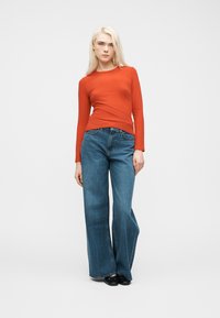 Νεαρή γυναίκα που στέκεται φορώντας πορτοκαλί μπλούζα με μακριά μανίκια, μπλε τζιν παντελόνια με φαρδιά πόδια και μαύρα παπούτσια, σε απλό λευκό φόντο.