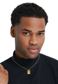 Collier en pendentif en or avec un charme rectangulaire, présentant un texte gravé. Porté par-dessus un haut noir à col montant, avec un design de chaîne lisse.