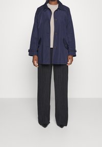 Trench coat blu navy con chiusura frontale a bottoni, ampie mostrine e tasche laterali, abbinato a pantaloni neri a pieghe a gamba larga e scarpe nere appuntite.