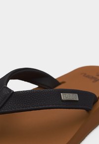 Καφέ σανδάλι σόλα με μαύρα υφασμάτινα λουριά και μικρή μεταλλική ετικέτα χαραγμένη με το όνομα της μάρκας "REEF" σε ένα λουρί.