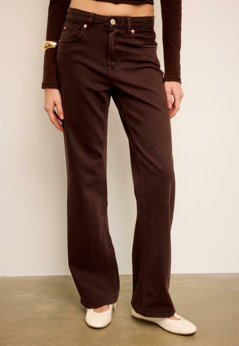 Femme portant un jean large taille haute couleur marron foncé, un haut court à manches longues, un bracelet en or et des ballerines blanches, debout sur un sol en béton.