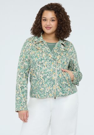 Vrouw met krullend haar, gekleed in een groen-geel patroonvest met ritssluiting, over een groene top en witte broek, poserend met één hand in haar zak.