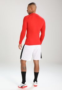 Rød langermet treningsskjorte med en figurtilpasset design, sammen med hvite shorts med svarte sidelister, iført svarte sokker og fotballsko.