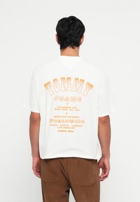White oversized t-shirt featuring orange text on the back, including "TOMMY JEANS" and various city names. Short sleeves, relaxed fit.