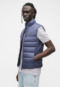 Vestă puff din bleumarin cu design matlasat, guler înalt și închidere cu fermoar, asortată cu un tricou gri deschis și blugi albastru deschis.